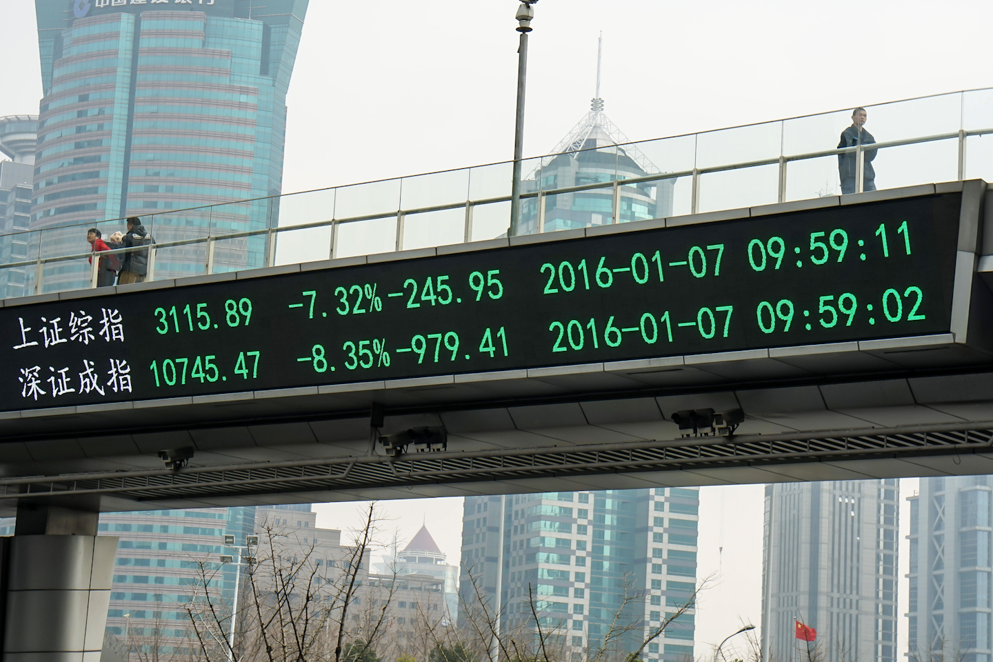 People walk across a pedestrian bridge with an electronic display showing figures for the Shanghai, top, and Shenzhen, bottom, stock exchanges in the Lujiazui Financial Zone in Shanghai, China. (Chinatopix via AP)