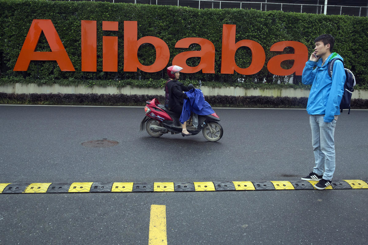 In this Friday, May 27, 2016, file photo, a man talks on his phone as a woman rides on an electric bike past a company logo at the Alibaba Group headquarters in Hangzhou in eastern China's Zhejiang province. Ford Motor Co. is collaborating with Chinese e-commerce giant Alibaba Group to further expand into the world&rsquo;s largest auto market. The carmaker signed a three-year agreement that will investigate ways to use technology for marketing, sales, cloud computing, and distribution strategies. It hopes to better incorporate digital technologies and platforms into its vehicles. (AP Photo/Ng Han Guan, File)