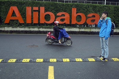In this Friday, May 27, 2016, file photo, a man talks on his phone as a woman rides on an electric bike past a company logo at the Alibaba Group headquarters in Hangzhou in eastern China's Zhejiang province. Ford Motor Co. is collaborating with Chinese e-commerce giant Alibaba Group to further expand into the world’s largest auto market. The carmaker signed a three-year agreement that will investigate ways to use technology for marketing, sales, cloud computing, and distribution strategies. It hopes to better incorporate digital technologies and platforms into its vehicles. (AP Photo/Ng Han Guan, File)