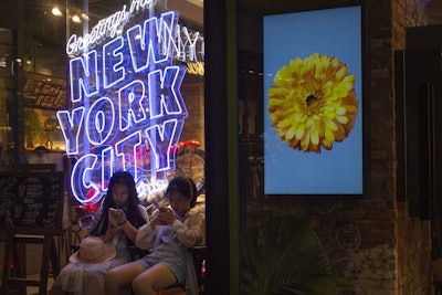 In this Thursday, July 5, 2018 photo, girls sit in front of an American cosmetics brand's shop window display reading 'Greetings from New York City' at a shopping mall in Beijing. Image credit: AP Photo/Mark Schiefelbein