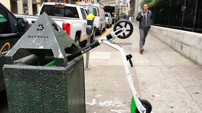 In this photo taken May 14, 2018, a man walks past an electric scooter that was dumped into a trash can in San Francisco. Tired of San Francisco streets being used as a testing ground for the latest delivery technology and transportation apps, city leaders are considering requiring businesses to get permits before trying out new high-tech ideas in public.