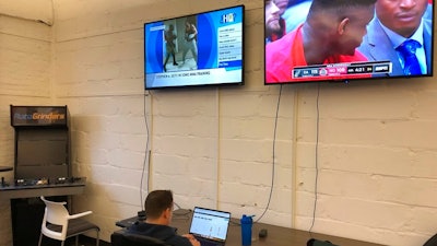 In this undated image provided by Cameron MacMillan, Brian Hourigan works at the Better Collective Tennessee, RotoGrinders.com office in downtown Nashville, Tenn., as NCAA college basketball plays on a television screen above. Sports can be a distraction at work the first few months of the year as pro football and college basketball hold their premier events. Business owners have to make a choice, whether, like McMillan at RotoGrinders.com, they'll allow or encourage staffers to enjoy the moment, or demand that everyone focus on work.