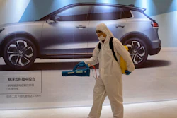 In this Feb. 12, 2020, photo, a worker disinfects a mall near an advertisement for a car in Beijing, China. Automakers are gradually reopening factories in China that were idled by anti-virus control.