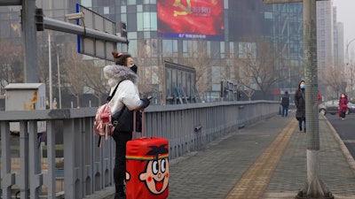 A traveler stands on a bridge near a display showing government propaganda in the fight against the COVID-19 viral illness in Beijing, China Thursday, Feb. 13, 2020. China is struggling to restart its economy after the annual Lunar New Year holiday was extended to try to keep people home and contain novel coronavirus. Traffic remained light in Beijing, and many people were still working at home.