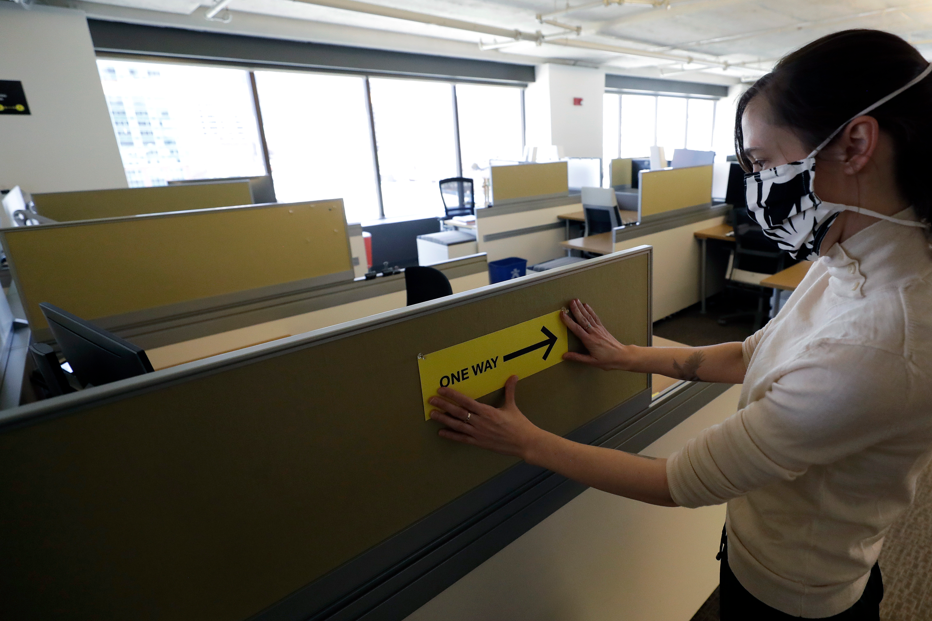 This Thursday, May 7 photo shows Interior Designer Stephanie Jones at the design firm Bergmeyer putting up a safe distancing reminder indicating one way foot traffic to a cubicle at the firms offices in Boston.