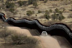 Customs and Border Control agent patrolling on the US side of a razor-wire-covered border wall.