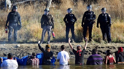In this Nov. 2, 2016 file photo, dozens of protestors demonstrating against the expansion of the Dakota Access Pipeline wade in cold creek waters confronting local police, near Cannon Ball, N.D. The Army Corps of Engineers is recommending that the federal government negotiate a settlement with North Dakota for more than $38 million that the state spent policing the monthslong pipeline protests against the Dakota Access oil pipeline almost four years ago.