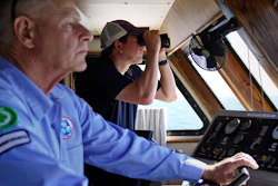 Aaron Smith, President and CEO of the Offshore Marine Service Association, center, peers through binoculars at ships installing the South Fork Wind project, as Capt. Rick Spaid, left, pilots the vessel Jones Act Enforcer, Tuesday, July 11, 2023, off the coast of Rhode Island.