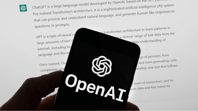 The OpenAI logo is seen on a mobile phone in front of a computer screen which displays output from ChatGPT, Tuesday, March 21, 2023, in Boston.