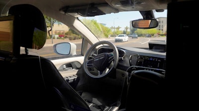 A Waymo minivan moves along a city street as an empty driver's seat and a moving steering wheel drive passengers during an autonomous vehicle ride, on April 7, 2021, in Chandler, Ariz.