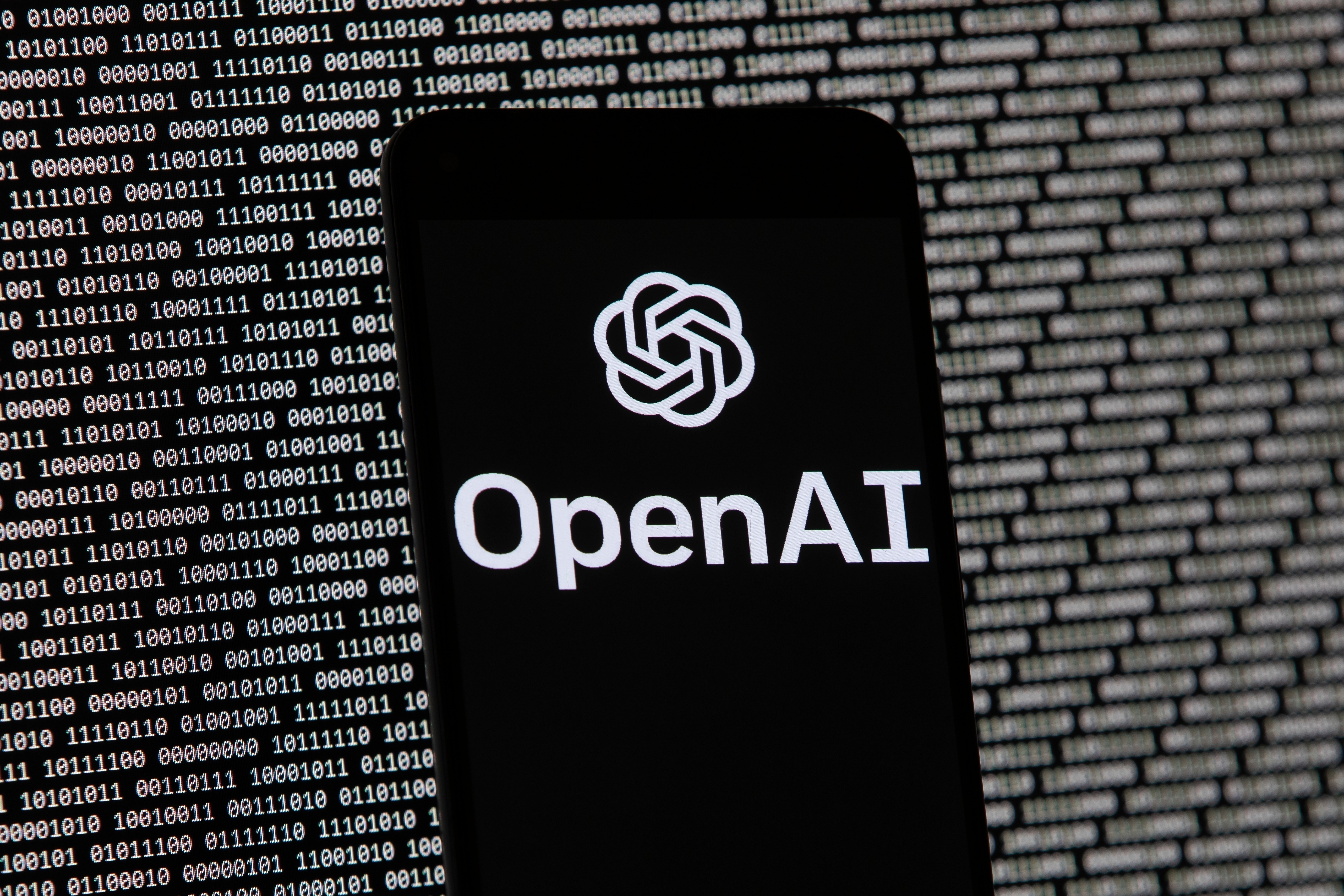 The OpenAI logo appears on a mobile phone in front of a computer screen with random binary data, March 9, 2023, in Boston.