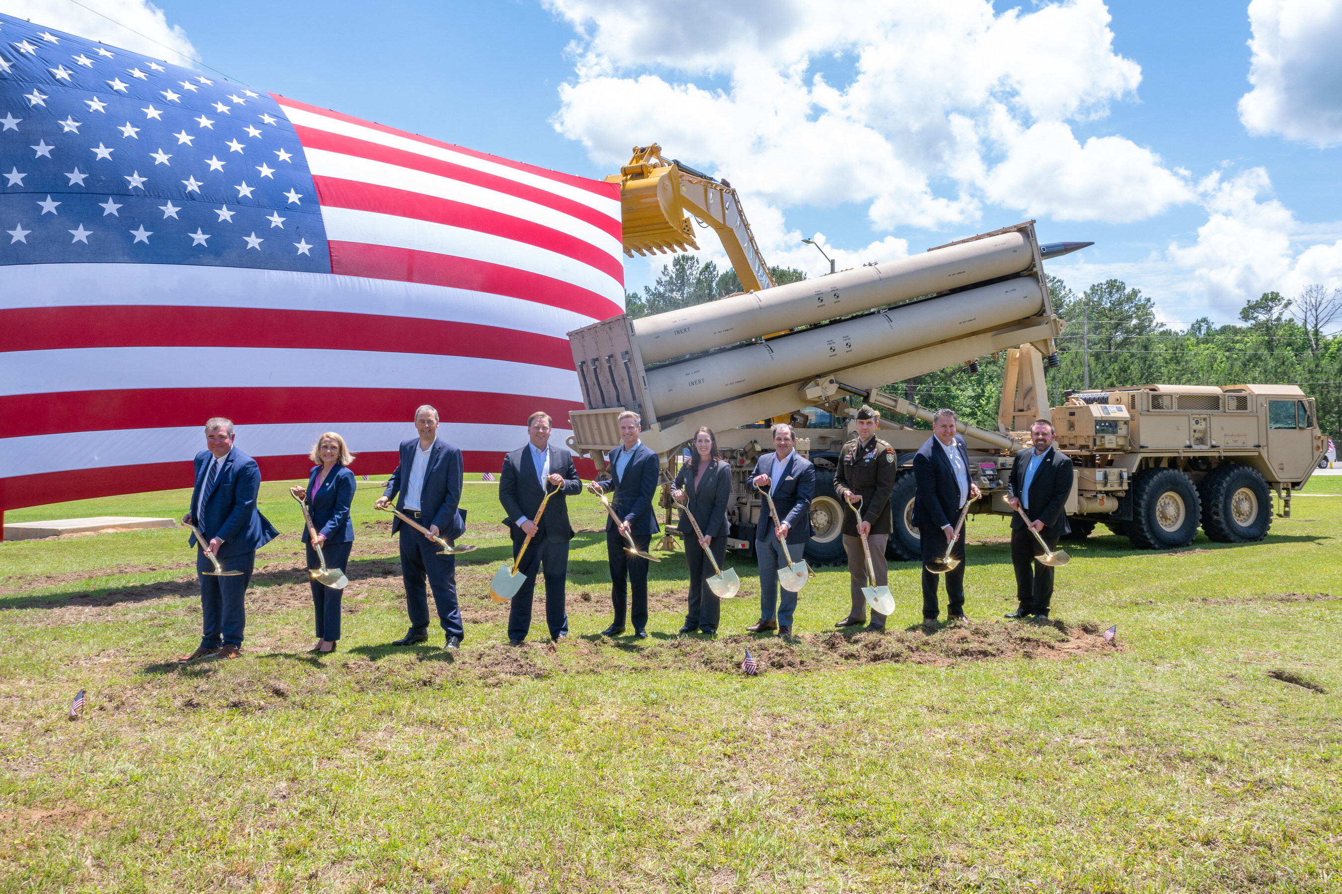 Military officials conduct groundbreaking ceremony with shovels before American flag and equipment