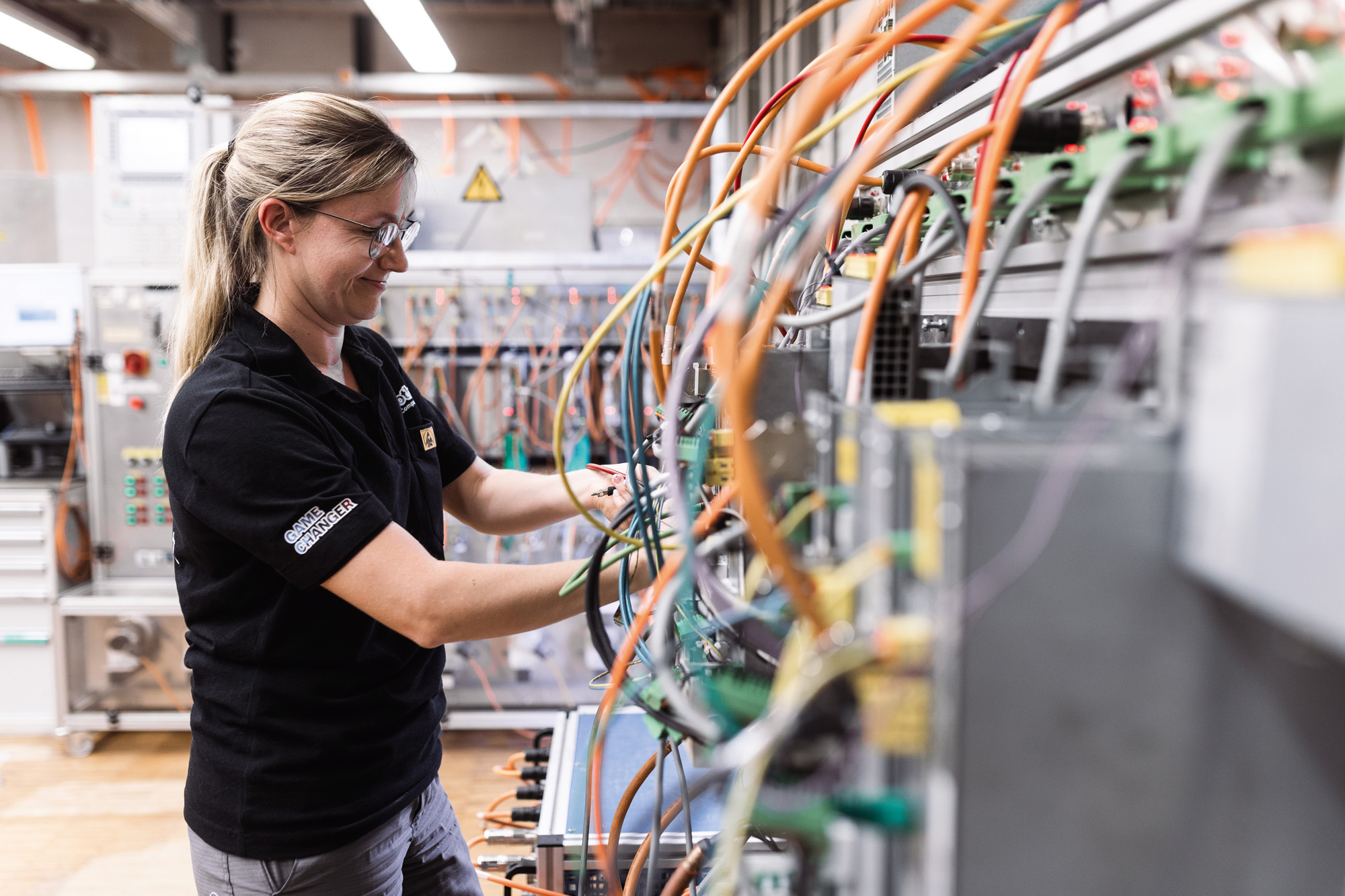 Female technician in safety glasses working on electrical panel with orange and blue wiring in industrial facility