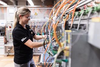 Female technician in safety glasses working on electrical panel with orange and blue wiring in industrial facility