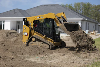 Yellow CAT 259 compact track loader moving soil at residential construction site with white house in background