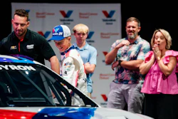 Racing team members and officials gather around a red and white race car during an automotive event presentation.