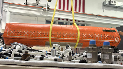 Orange cylindrical aerospace equipment suspended by yellow straps in industrial workshop with American flags