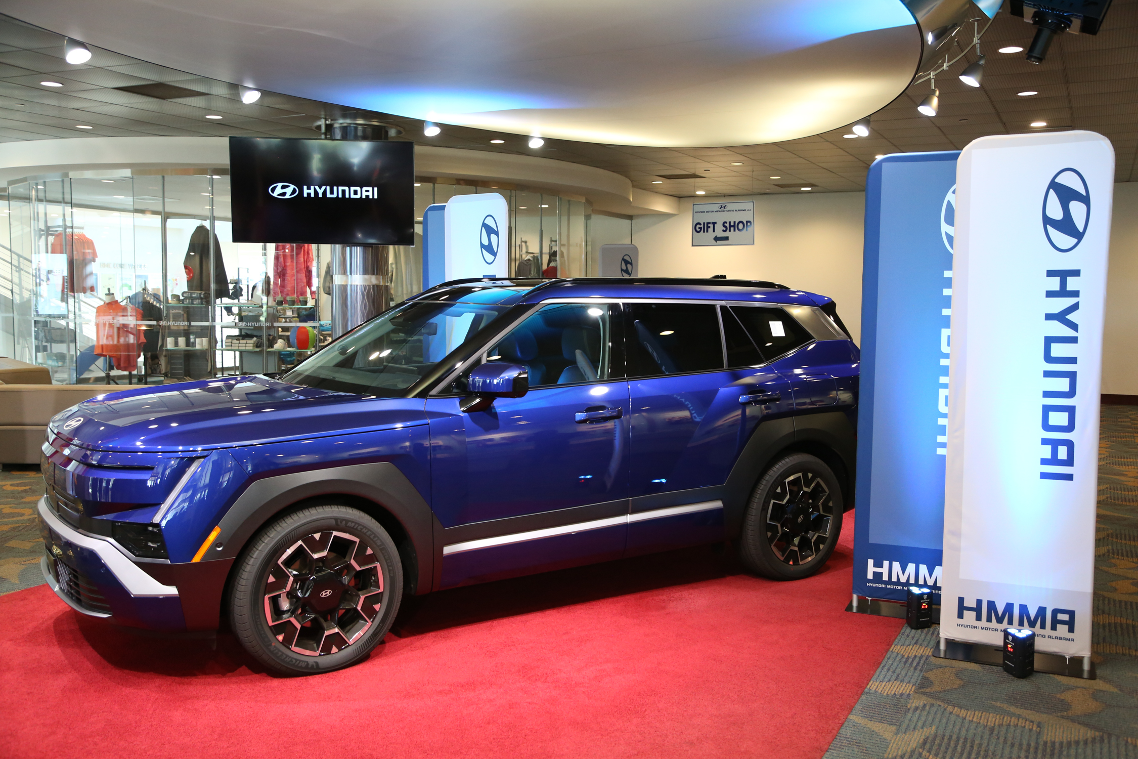 Blue Hyundai Kona electric vehicle displayed in modern showroom with branded signage and red carpet