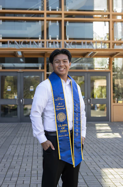 Smiling graduate in white shirt wearing blue and gold UC Irvine Electrical Engineering Class of 2026 honor stole outside engineering building