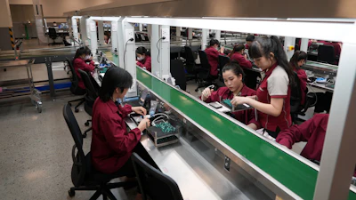 Electronics factory workers in maroon uniforms assembling components at industrial workstations in a bright manufacturing facility.