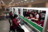 Electronics factory workers in maroon uniforms assembling components at industrial workstations in a bright manufacturing facility.