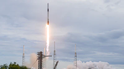 SpaceX Falcon 9 rocket launches from coastal launch pad with bright engine flames and white smoke clouds against cloudy sky