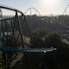 Aerial view of blue roller coaster tracks winding through an amusement park surrounded by trees and landscape