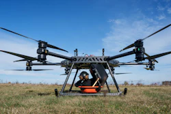 Large agricultural drone with orange payload container hovering above dry field under clear blue sky