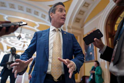 Man in blue suit and gold tie speaking to media in ornate government building corridor