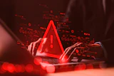 Red warning alert symbol glowing above laptop with hands, surrounded by digital data icons and circuit patterns on dark background