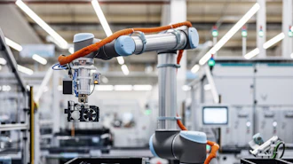 Two orange and silver robotic arms working together in a modern manufacturing facility with white structural elements and industrial equipment.