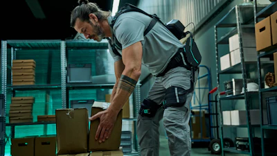 Worker in gray shirt and tool belt organizing cardboard boxes in a modern warehouse with teal shelving.