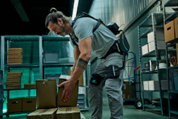 Worker in gray shirt and tool belt organizing cardboard boxes in a modern warehouse with teal shelving.