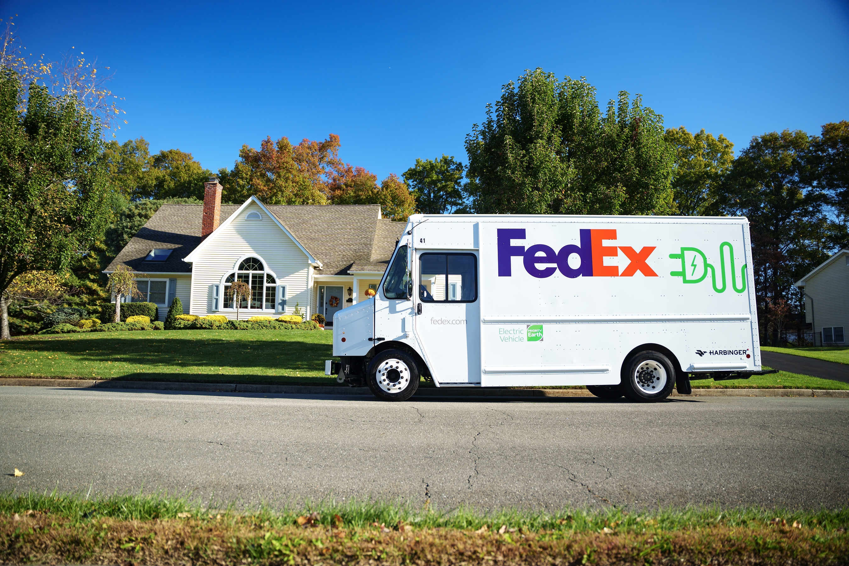 FedEx delivery truck parked on residential street in front of white house with autumn trees