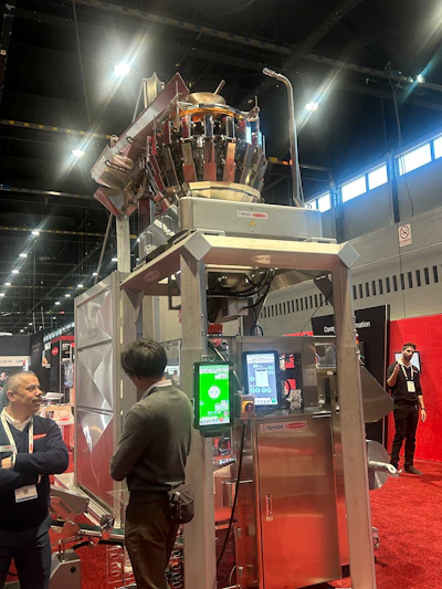 Industrial vertical form-fill-seal packaging machine on display at trade show with operator interface panel and red exhibition booth backdrop