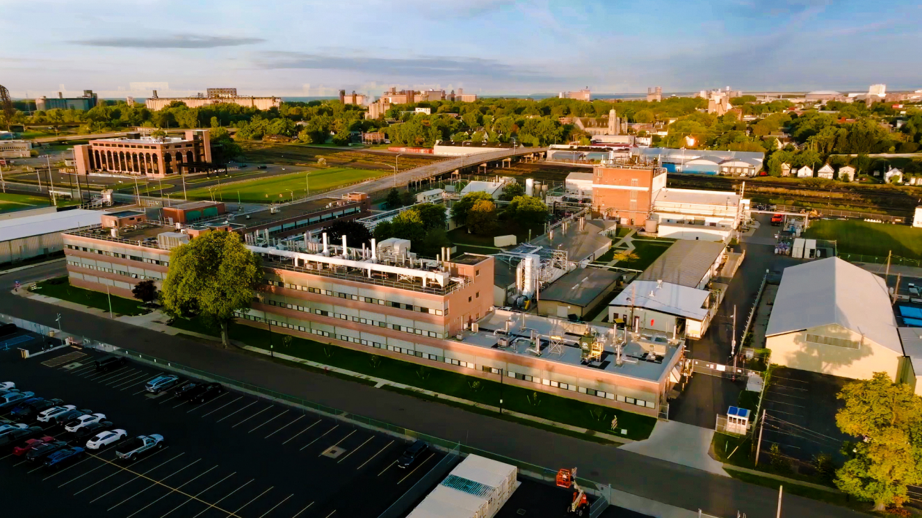 Aerial view of brick industrial complex with parking lot, green spaces, and surrounding cityscape