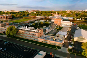 Aerial view of brick industrial complex with parking lot, green spaces, and surrounding cityscape