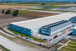Aerial view of modern industrial warehouse complex with blue-accented buildings, loading docks, and surrounding agricultural landscape.