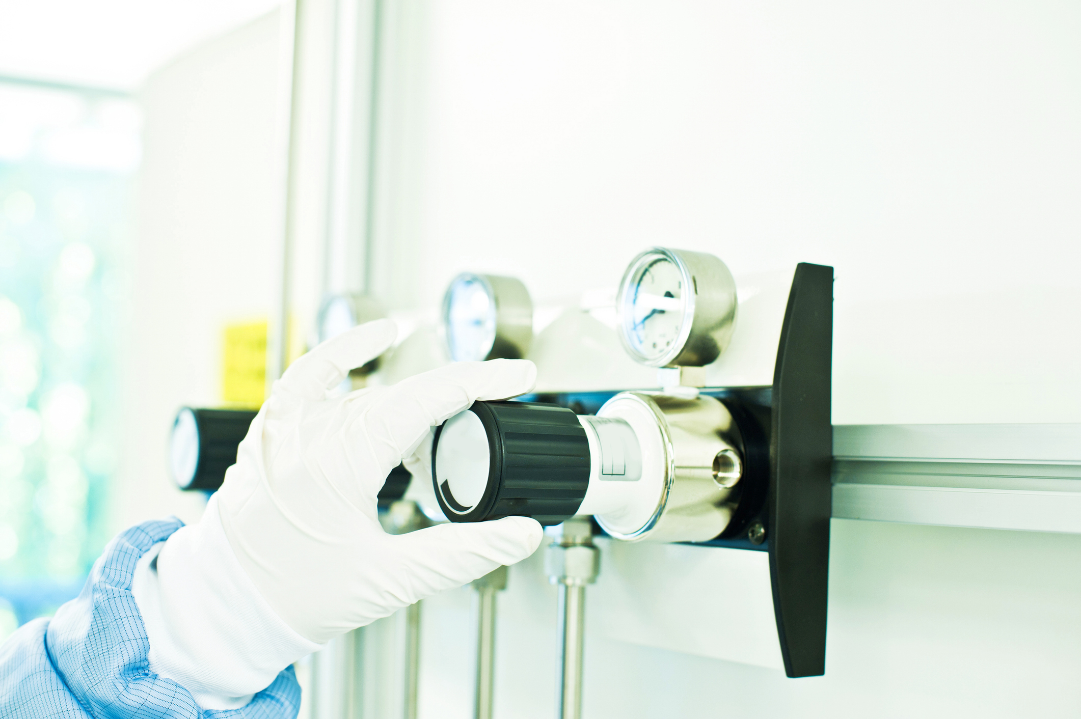 Gloved hand holding a brass gas regulator valve with pressure gauges and black control knob in laboratory setting