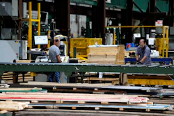 Workers processing lumber at an industrial woodworking facility with yellow machinery and green structural beams.