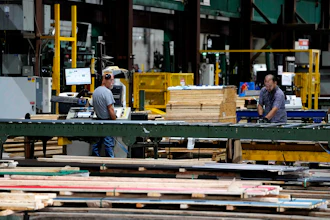 Workers processing lumber at an industrial woodworking facility with yellow machinery and green structural beams.