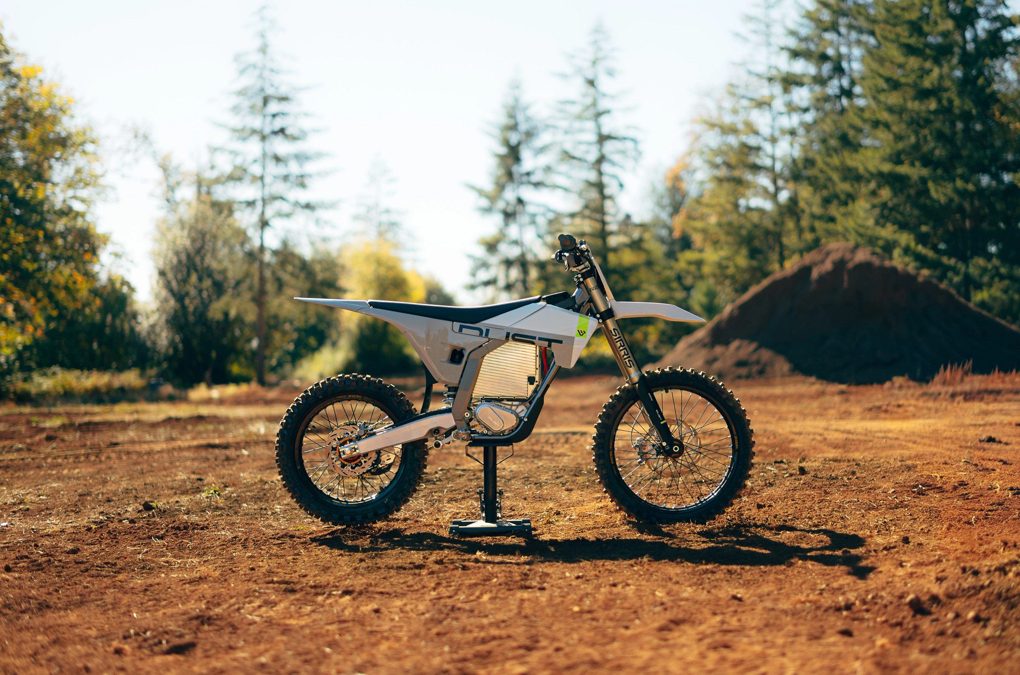 White electric dirt bike displayed on brown dirt ground with coniferous forest and autumn trees in background