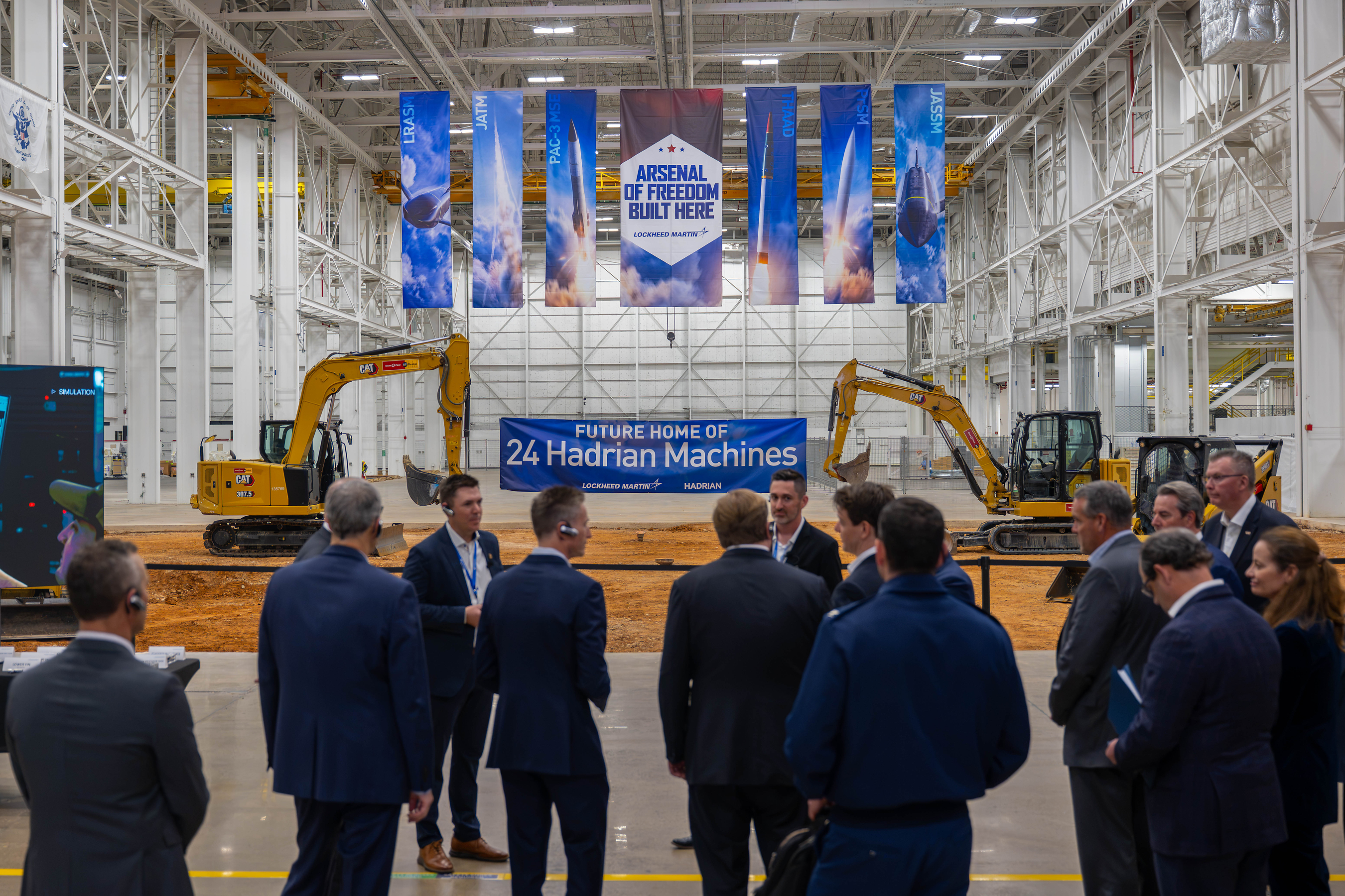 Corporate event in industrial warehouse with blue banner reading 'Future Home of 24 Hadrian Machines' and yellow excavators