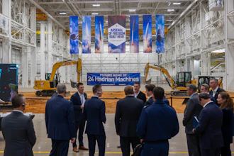 Corporate event in industrial warehouse with blue banner reading 'Future Home of 24 Hadrian Machines' and yellow excavators