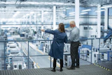 Two professionals observe a large industrial manufacturing facility from an elevated platform with expansive windows.