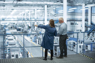 Two professionals observe a large industrial manufacturing facility from an elevated platform with expansive windows.