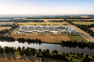 Aerial view of industrial facility with blue-roofed buildings, storage tanks, and surrounding agricultural fields near a river