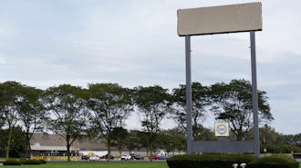 Large blank billboard with metal frame in commercial area surrounded by trees, buildings, and parked vehicles