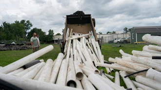 White PVC pipes being unloaded from a dump truck at an industrial yard with green buildings in background