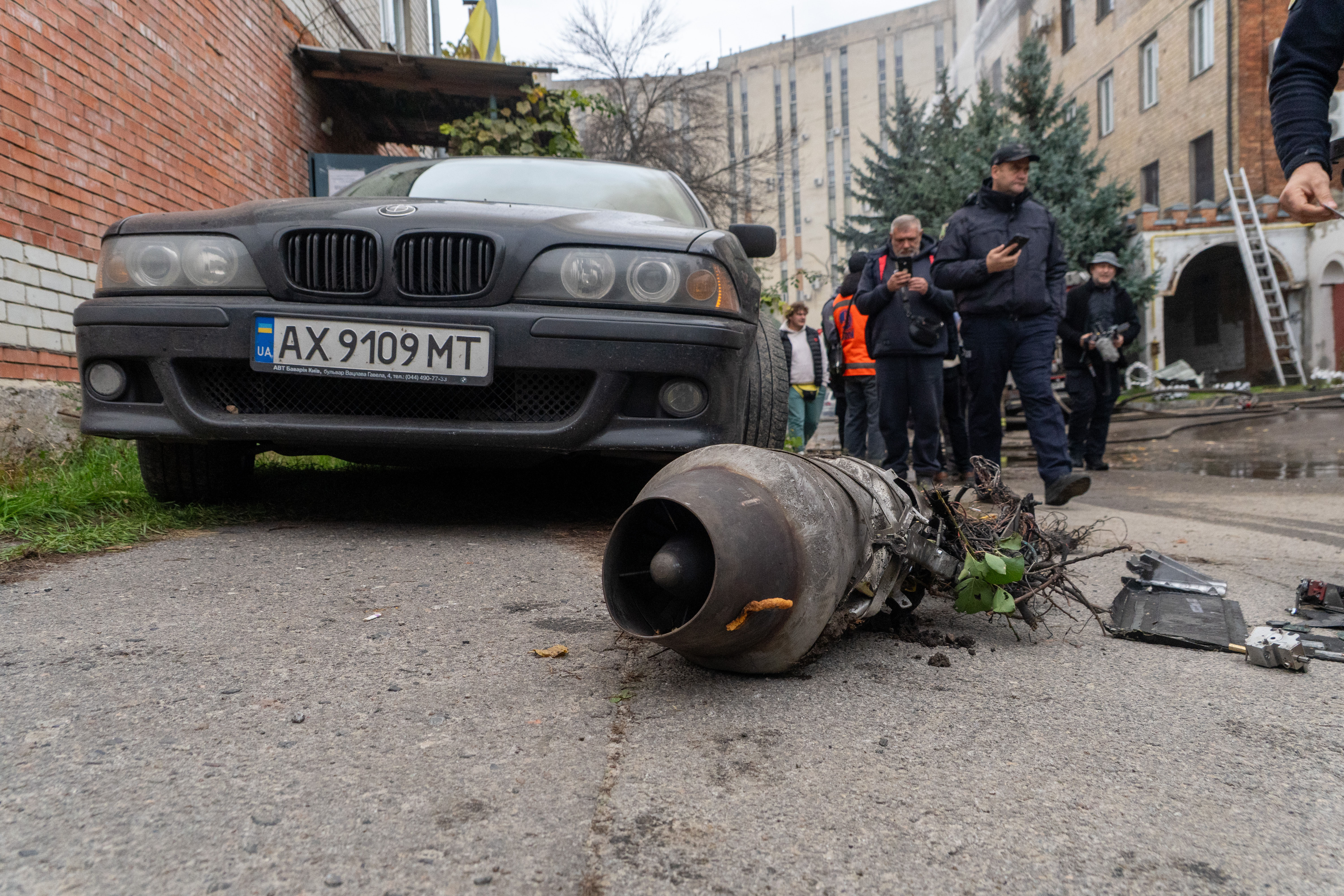 Damaged BMW sedan with detached engine component displayed on ground during investigation with officials and onlookers in urban courtyard